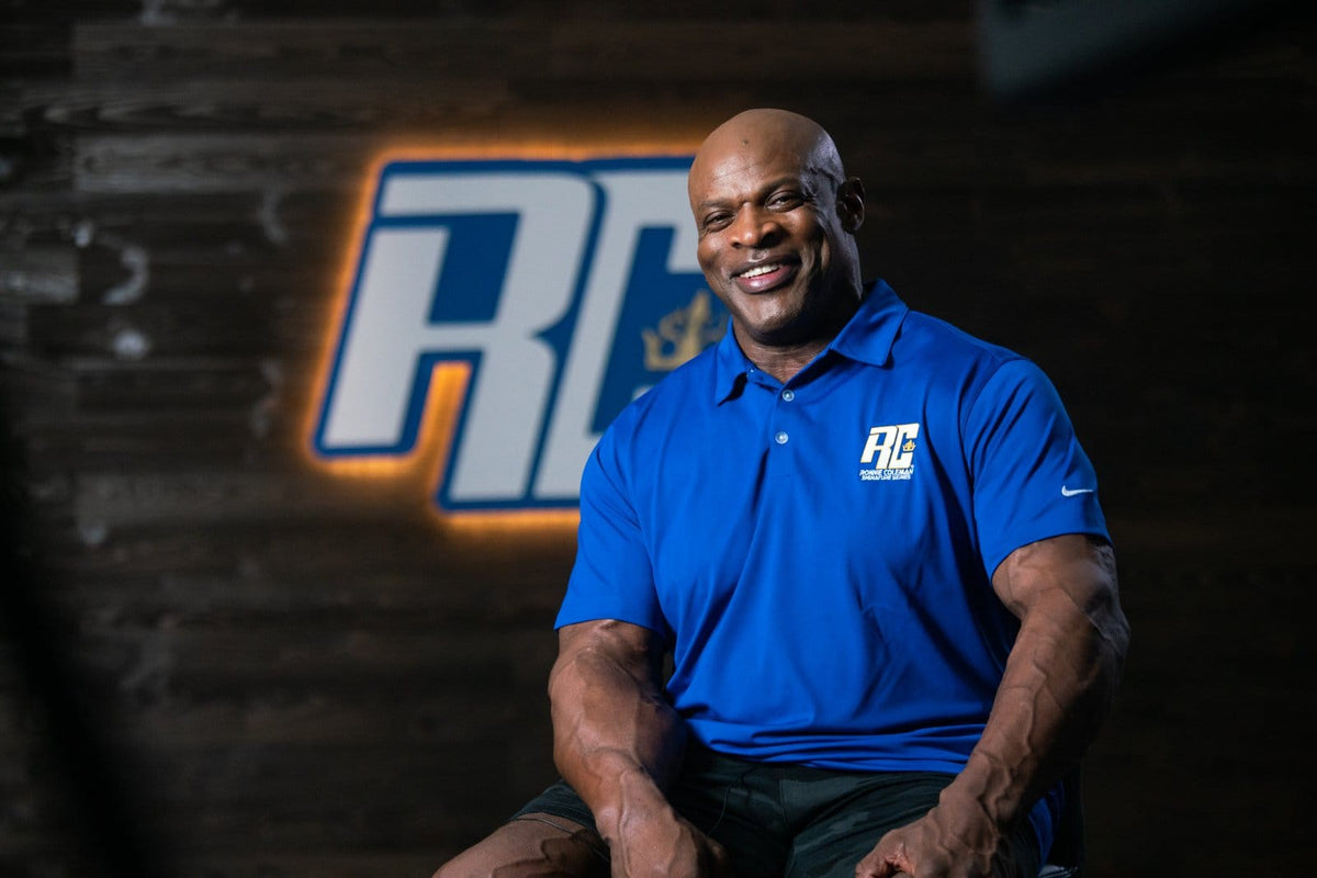Ronnie Coleman in a blue RC polo in front of the RC logo wall